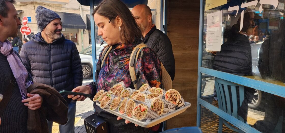 Person holding a tray of wrapped sandwiches on a busy street.