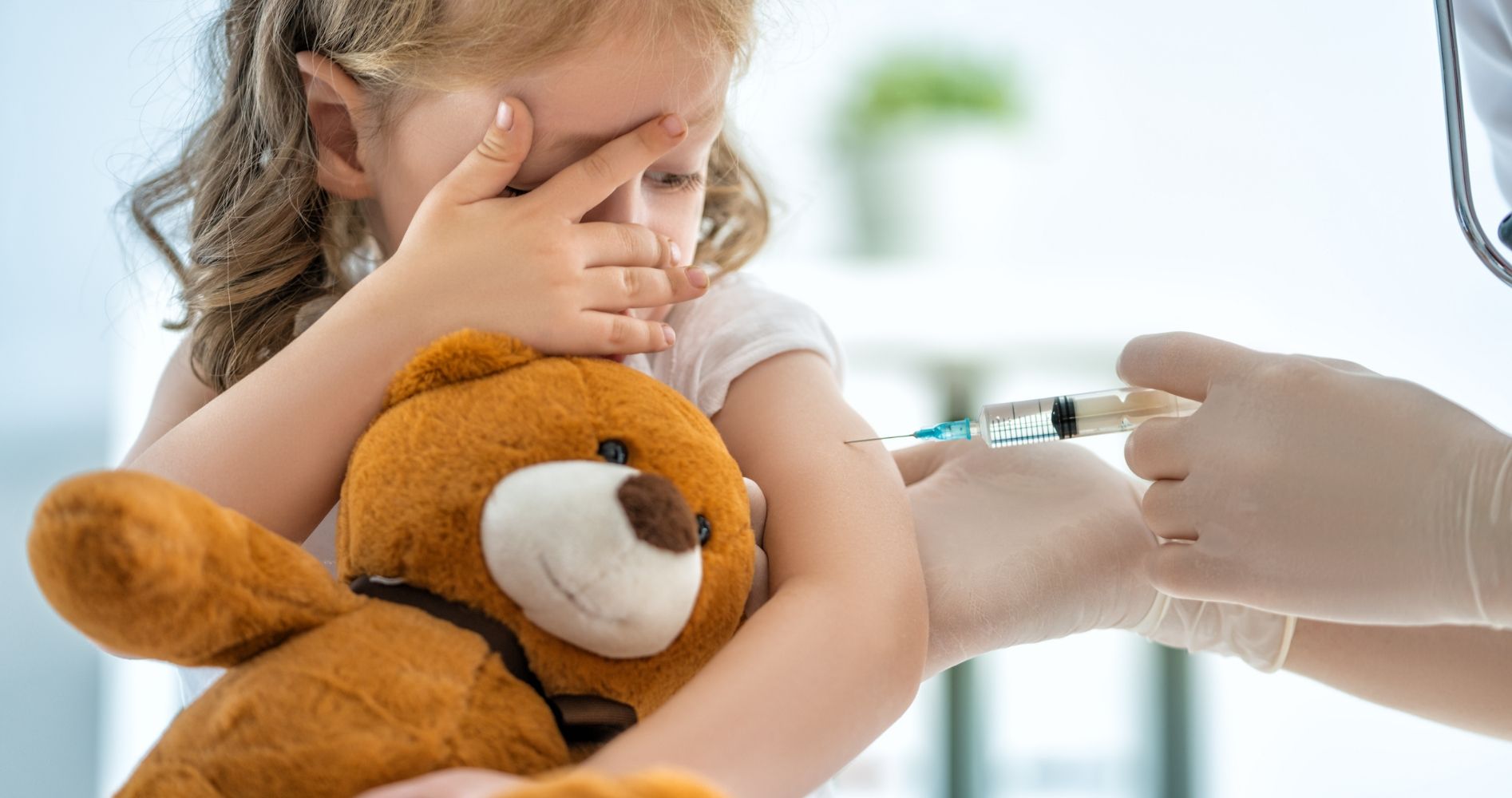 Child holding teddy bear while a doctor prepares to give a vaccine.